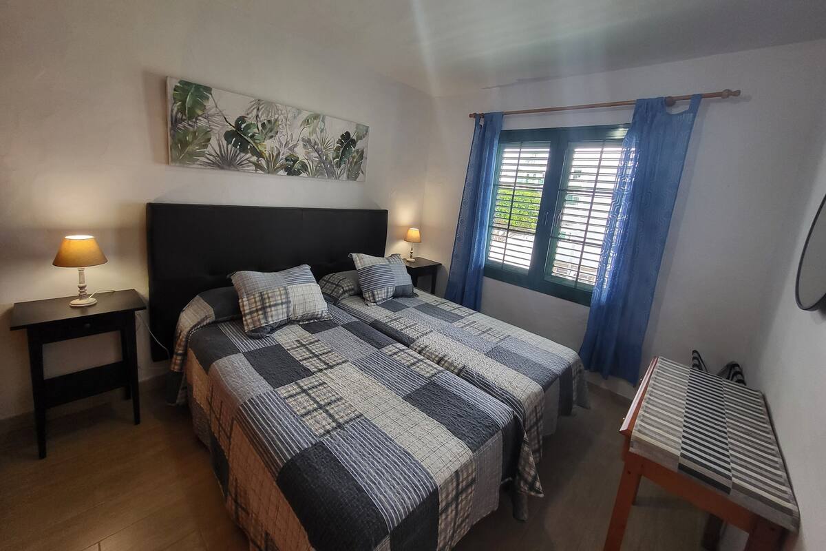 A bedroom with a large bed, two bedside tables, a wall-mounted lamp, a window with curtains, and a painting on the wall.