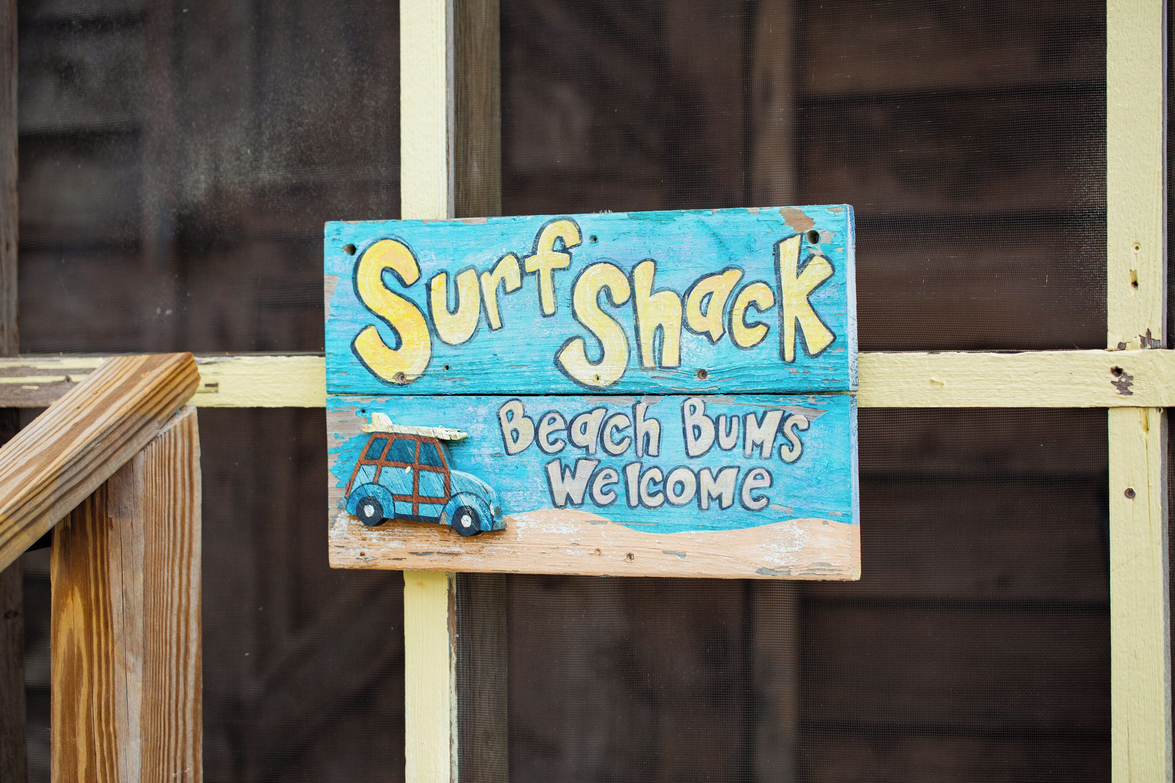 A wooden sign with the text "Surf Shack Beach Bums Welcome" and a drawing of a vintage car.