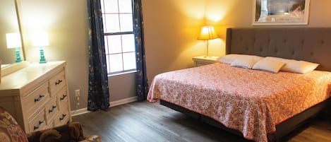 Un dormitorio con cama, cómoda, silla, ventana con cortinas, lámpara de noche y un cuadro en la pared.