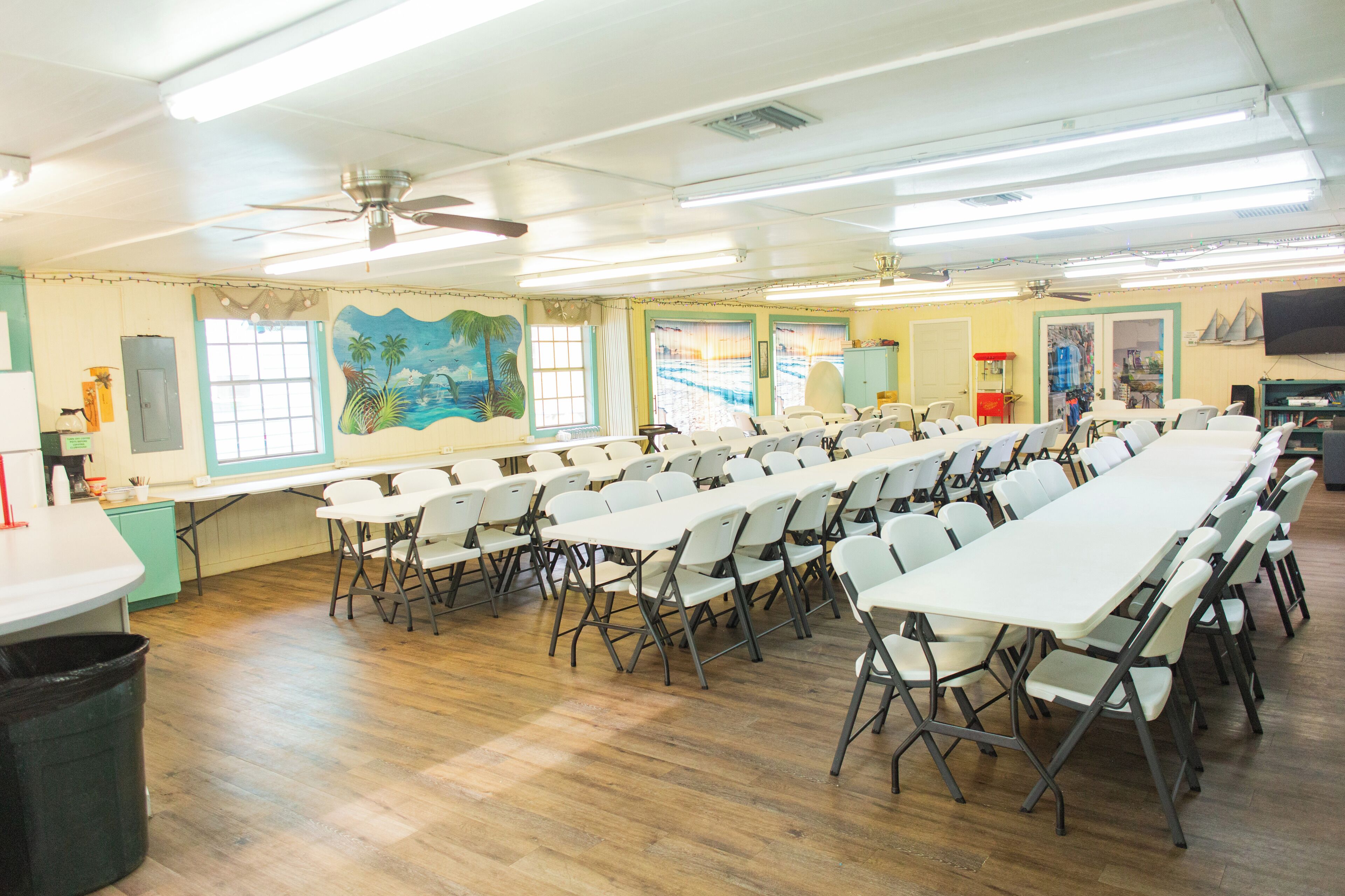 Une salle de conférence avec des tables et des chaises blanches, un ventilateur de plafond et des fenêtres ornées de peintures murales aux motifs tropicaux.