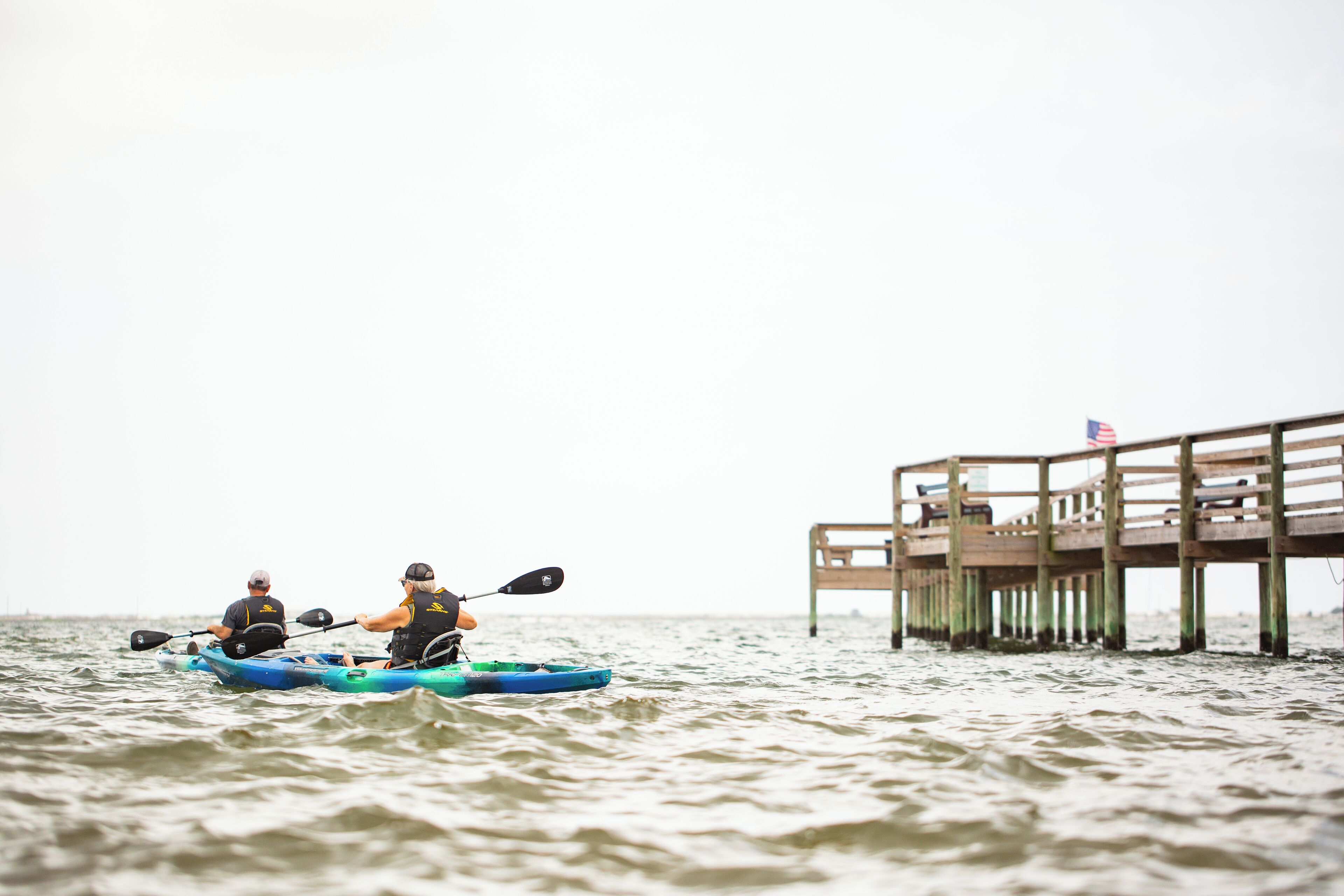 Dos personas en kayak sobre el agua, con un muelle de madera al fondo.