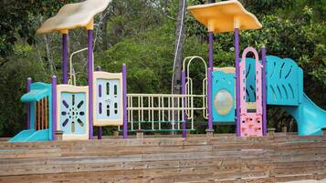 Una terraza de madera con un parque infantil que incluye toboganes, zonas de escalada y un espacio pequeño cerrado.