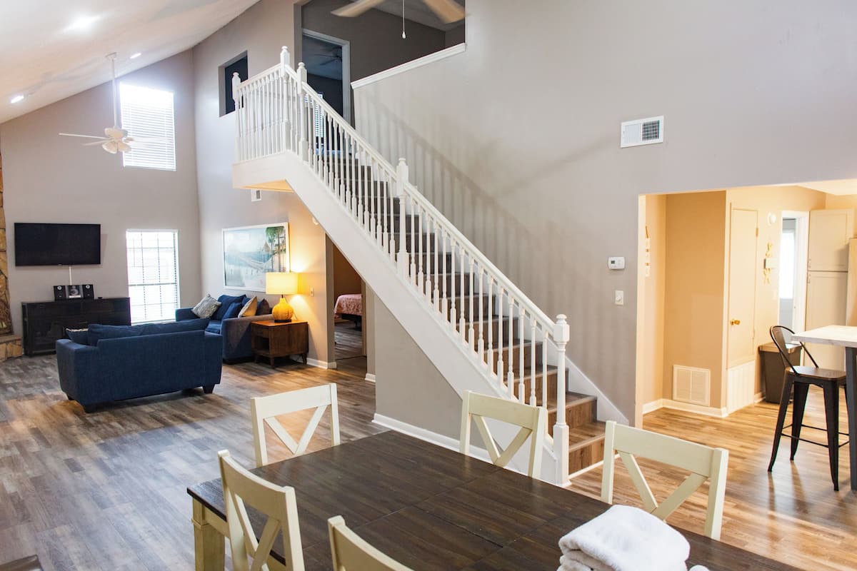Un vaste espace de vie doté d’un plancher en bois, d’une table à manger avec des chaises, d’un escalier avec des rampes blanches et d’un téléviseur à écran plat fixé au mur.