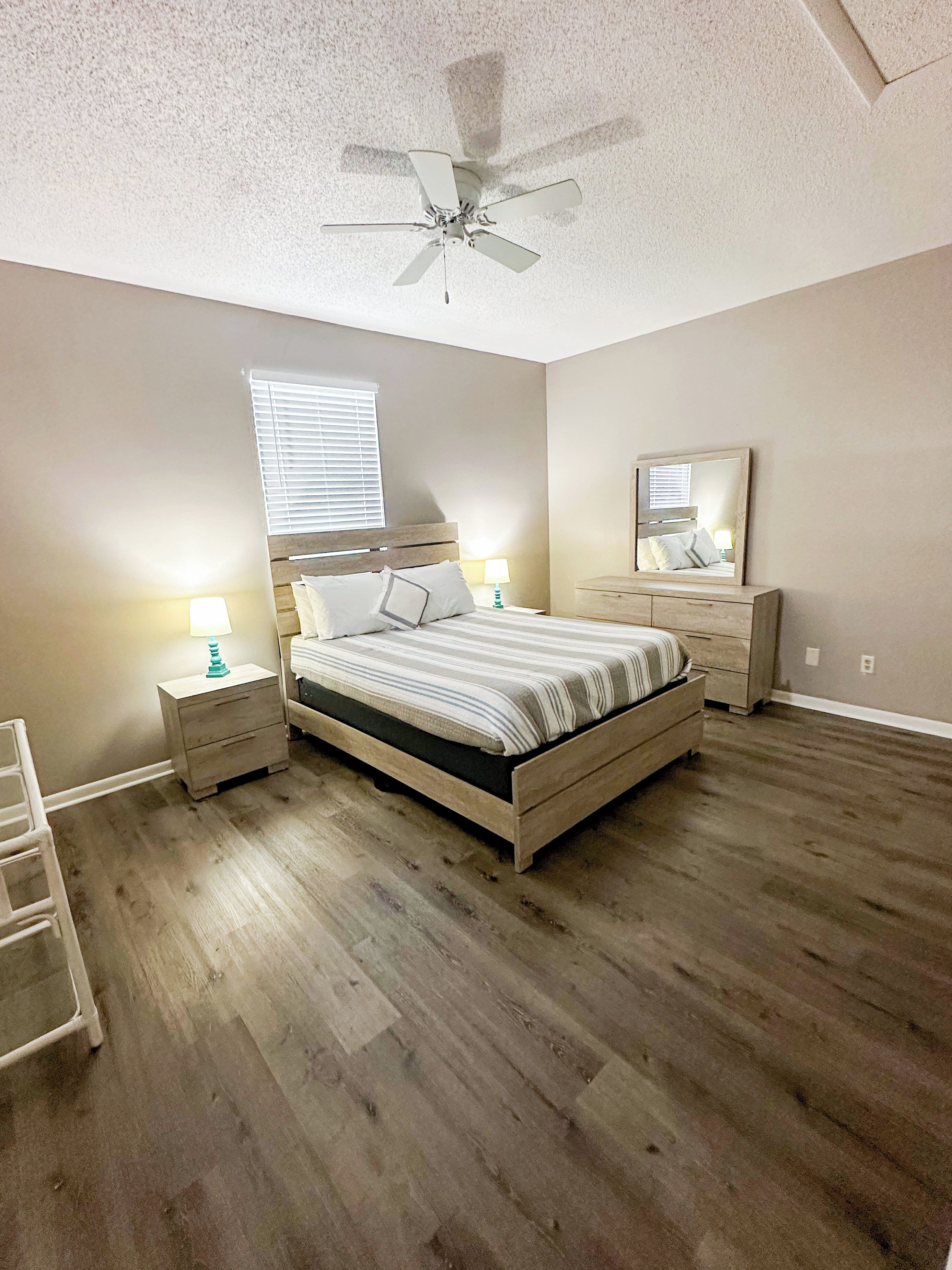 A bedroom with a wooden bed, nightstands, a ceiling fan, a window with blinds, a mirror, and a wooden floor.
