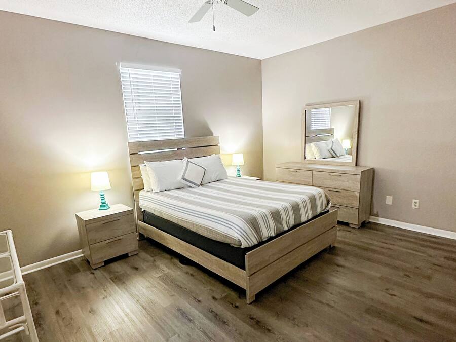 Een slaapkamer met een houten bed, nachtkastjes, een plafondventilator, een raam met jaloezieën, een spiegel en een houten vloer.