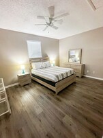 A bedroom with a wooden bed, nightstands, a ceiling fan, a window with blinds, a mirror, and a wooden floor.