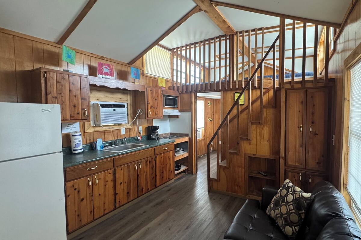 L’intérieur d’un chalet rustique, avec des murs en bois, une cuisine équipée d’un réfrigérateur, d’une cuisinière et d’un évier, un escalier et un espace de vie comprenant un canapé.