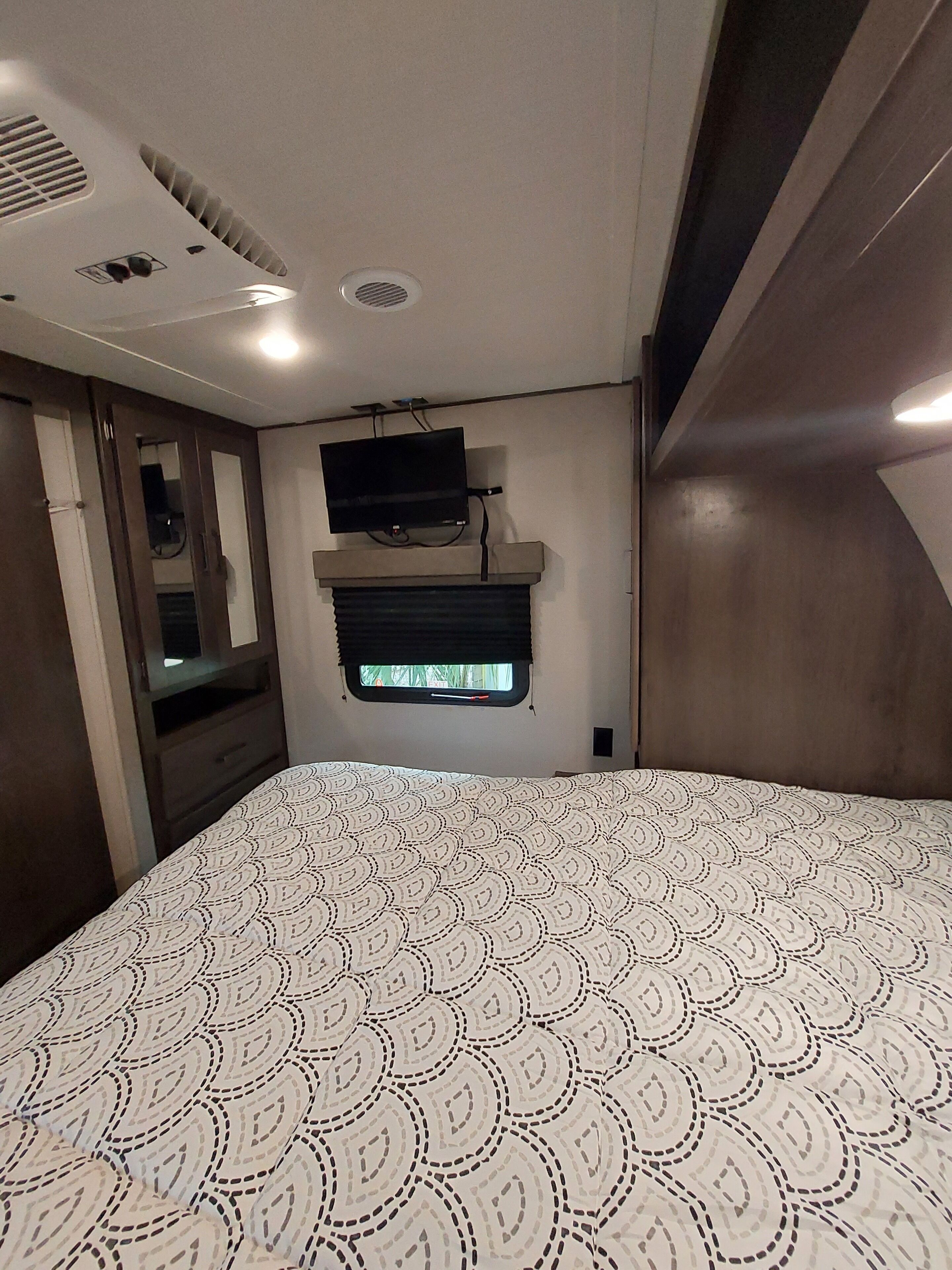 A bedroom with a patterned bedspread, a mounted television, and a window with blinds.