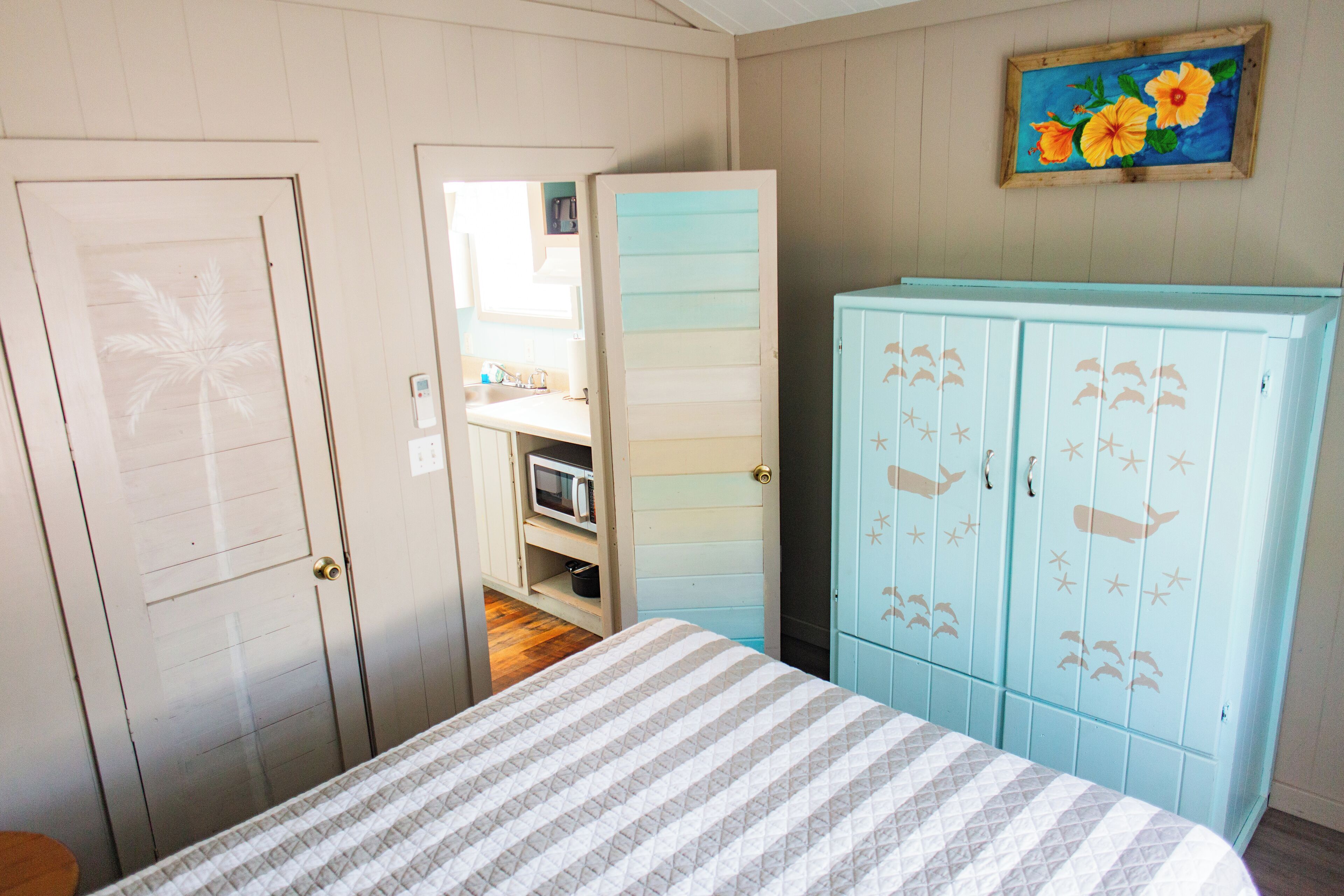 Une chambre avec un lit, une armoire décorée de motifs représentant des créatures marines, une porte ornée d’un motif de palmier et un tableau de fleurs accroché au mur.
