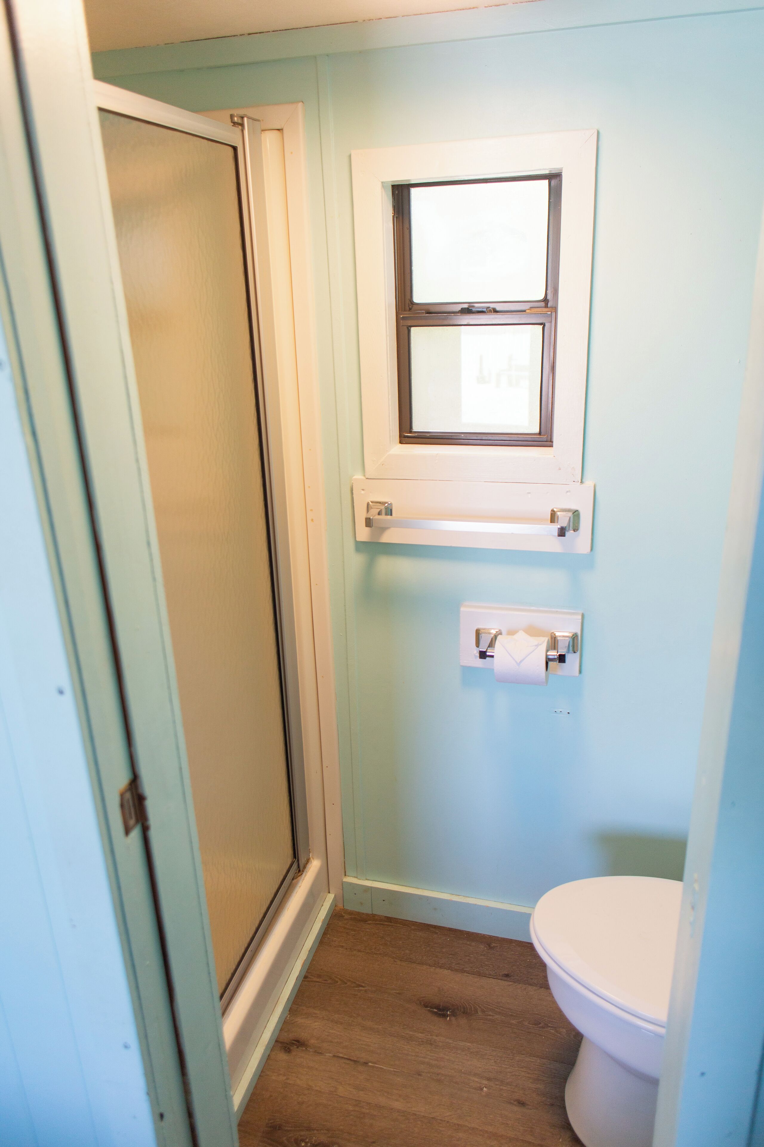 Et lille badeværelse med trægulv, et hvidt toilet, et vindue over vasken og en håndklædeholder.