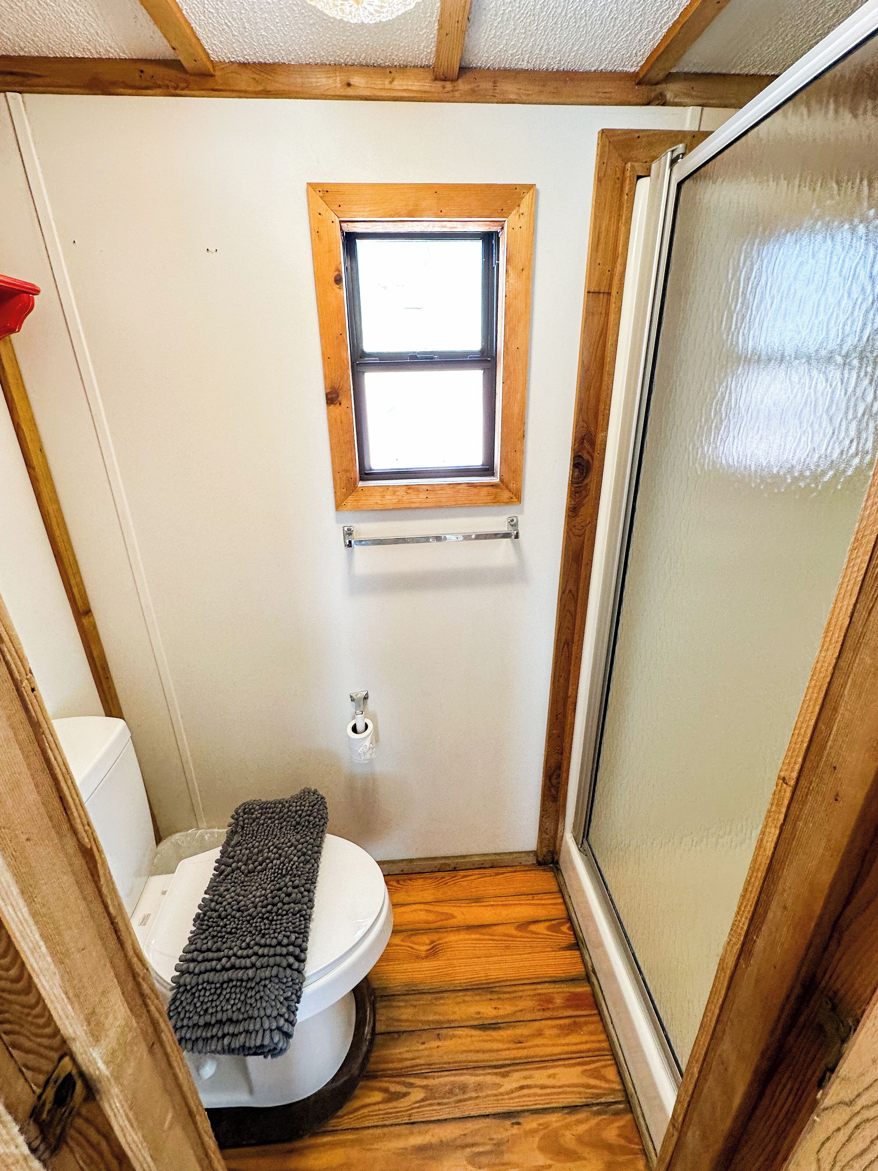 Une petite salle de bain avec un plancher en bois, des toilettes blanches, une petite fenêtre et un espace douche.