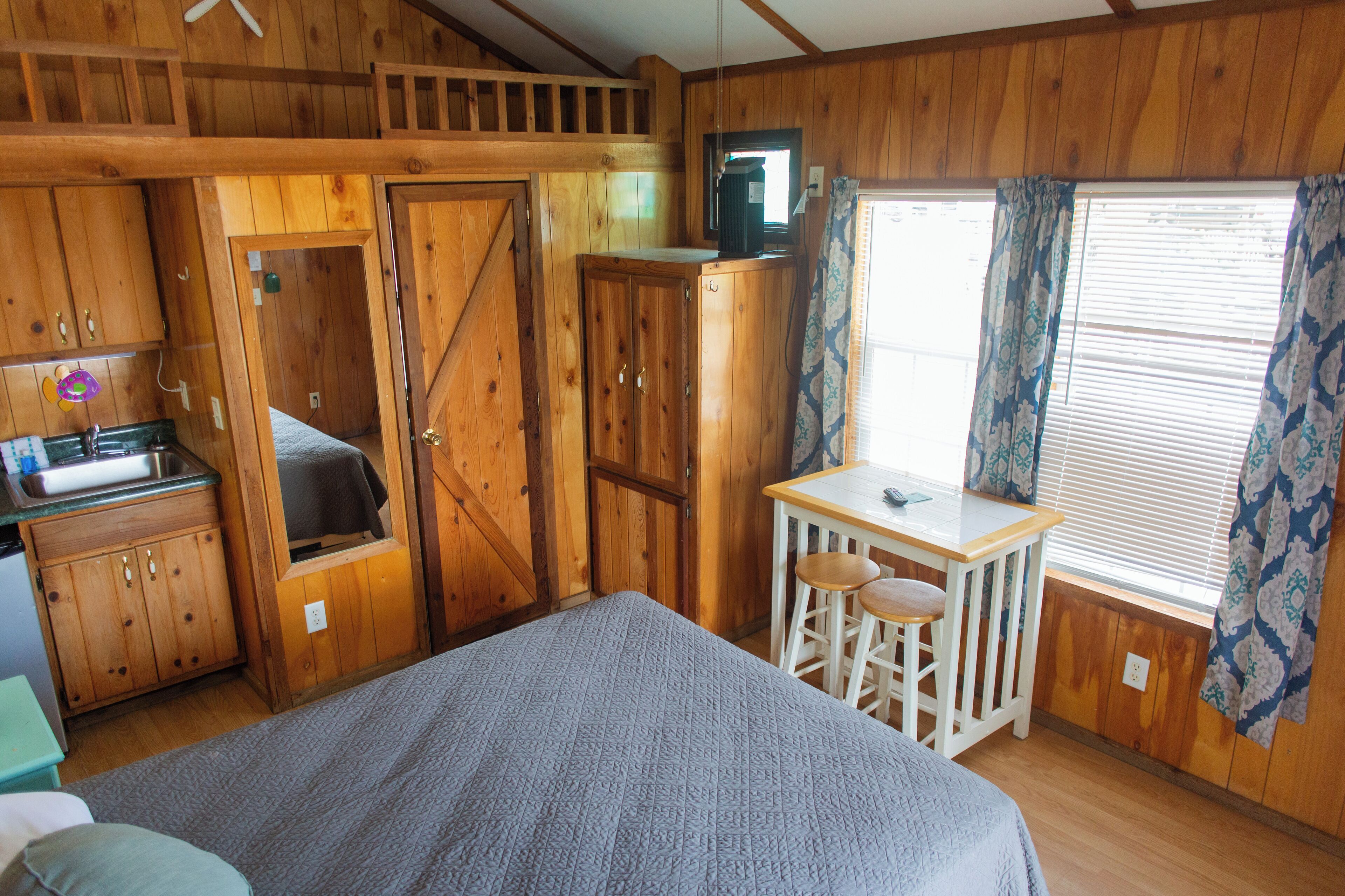 A wooden cabin room with a bed, a small table with two stools, a kitchenette, and a window with curtains.