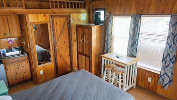 A wooden cabin room with a bed, a small table with two stools, a kitchenette, and a window with curtains.