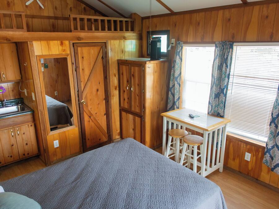 A wooden cabin room with a bed, a small table with two stools, a kitchenette, and a window with curtains.
