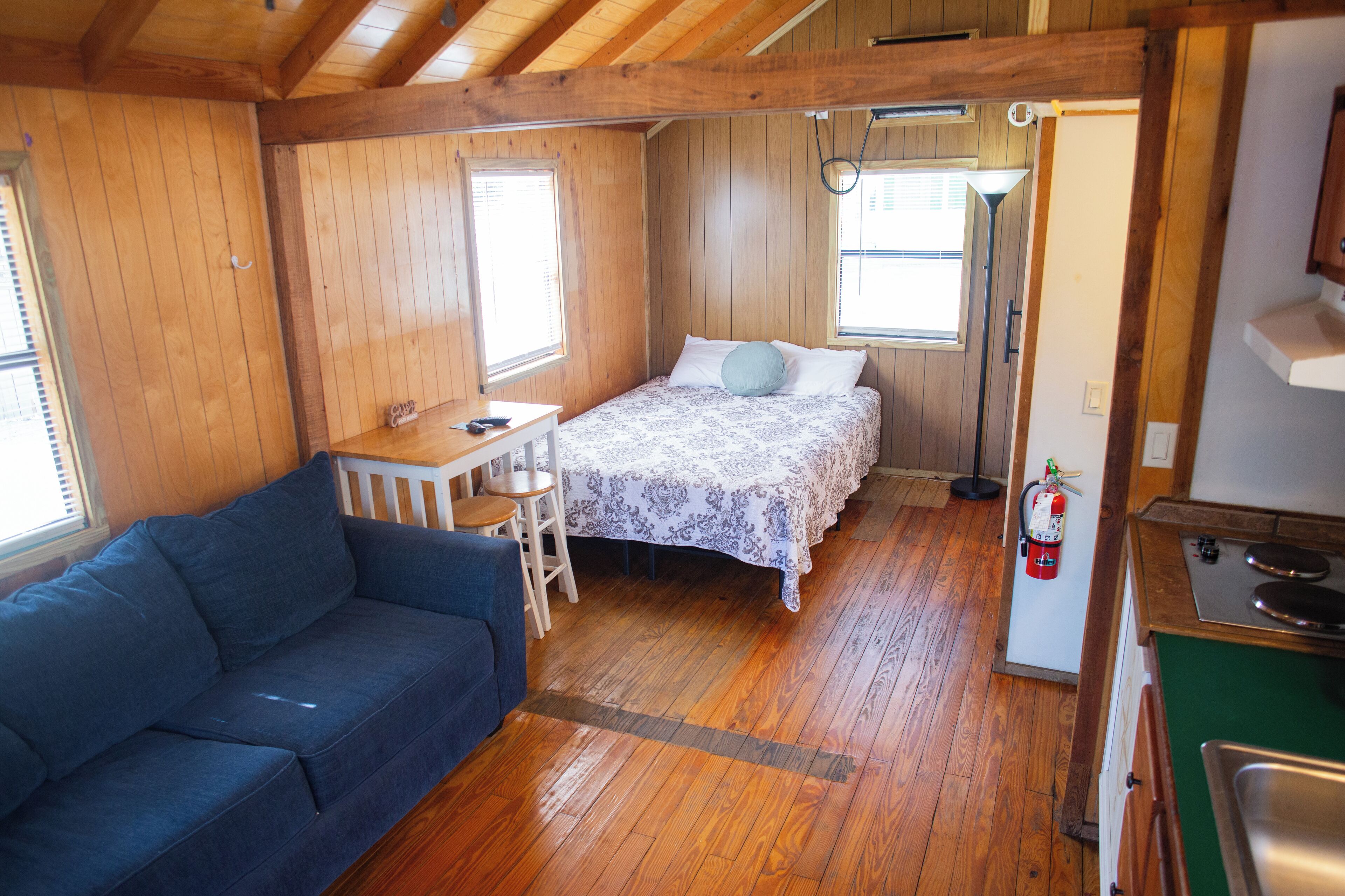 Una cabaña acogedora con interior de madera, una cama con colcha estampada, un sofá azul, una mesita y una zona de kitchenette.