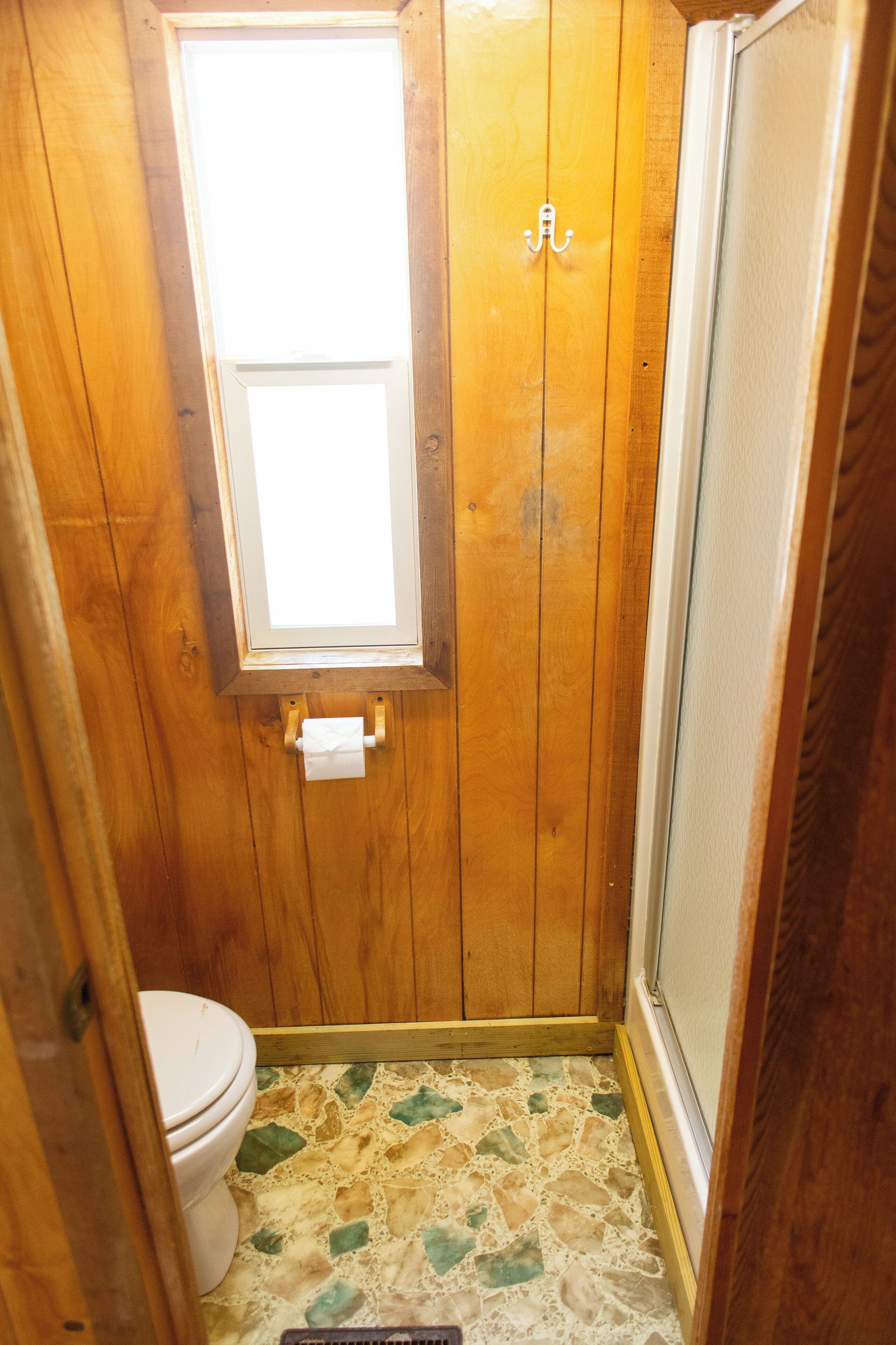 A small bathroom with wooden walls, a toilet, a window, and a shower.