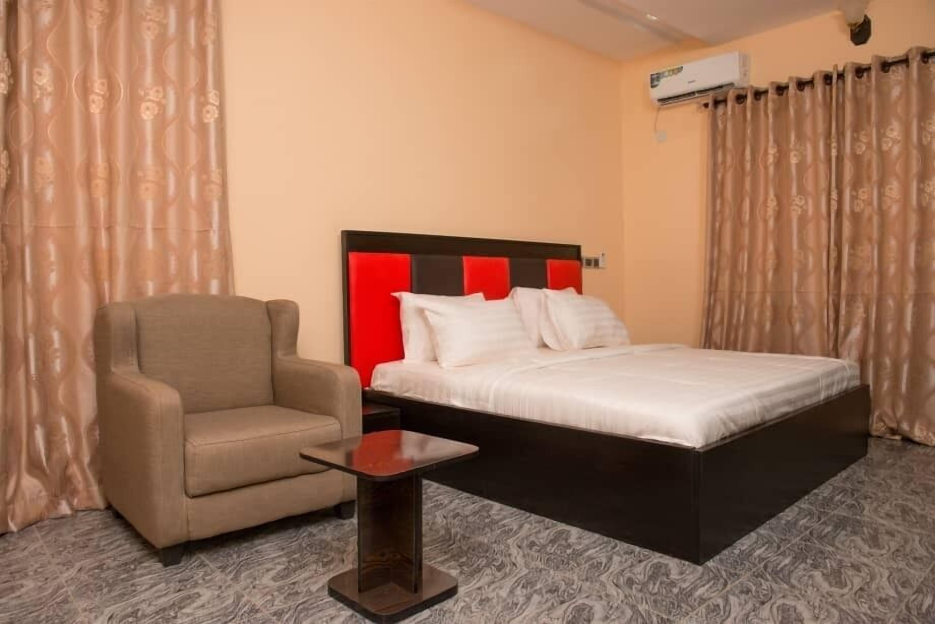 A hotel room with a bed, a chair, a small table, and an air conditioning unit.