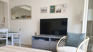 A living room with a flat-screen TV, a dining table, chairs, a wicker chair with a cushion, and wall art.
