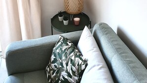 A living room with a grey sofa, a small round table with a woven lamp, two potted plants, and a framed artwork on the wall.