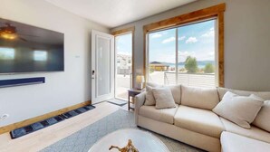 Un salon moderne doté d’un grand téléviseur à écran plat, d’un canapé beige et offrant une vue sur l’extérieur grâce à de grandes fenêtres.
