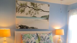 Un dormitorio con una cama, dos lámparas de noche, un ventilador de techo y un cuadro en la pared que muestra una playa.