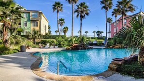 Ein Swimmingpool, umgeben von Palmen und Grünpflanzen, mit einem Gebäude im Hintergrund.