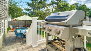 A barbecue grill on a wooden deck with a pool and patio furniture in the background. - Walk to Haigis Beach: Cape Cod Retreat w/ Deck! (Dennis)