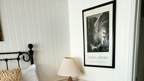A neatly made bed with a brown headboard, a red quilt, and patterned pillows. A framed picture of a forest scene hangs on the wall above the bed.