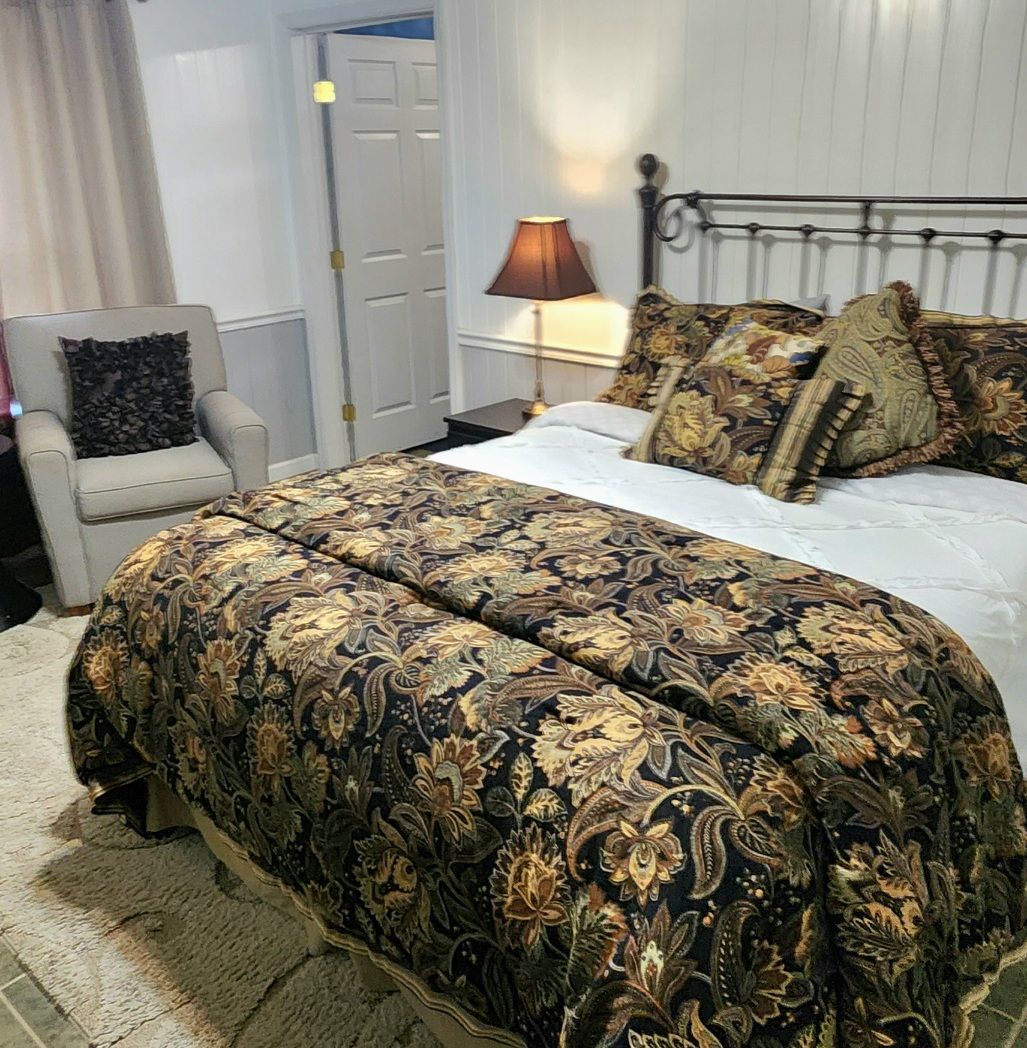 A bedroom with a large bed, a bedside table with a lamp, a chair, and a door in the background.