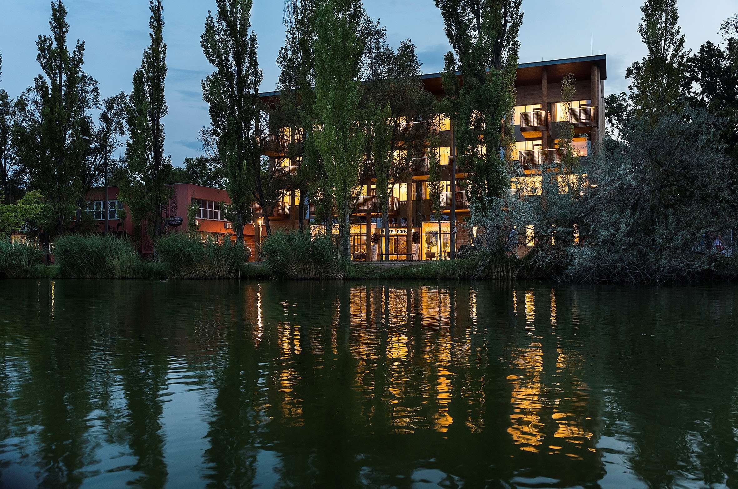 A multi-story hotel with balconies, a lake in the foreground, and trees around.