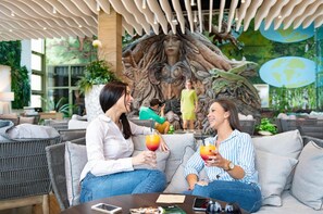 A lounge area with wicker furniture, a mural with a tree and human figures, and a glass table with drinks.