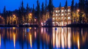 A multi-story hotel with balconies, a lake in the foreground, and trees in the background.
