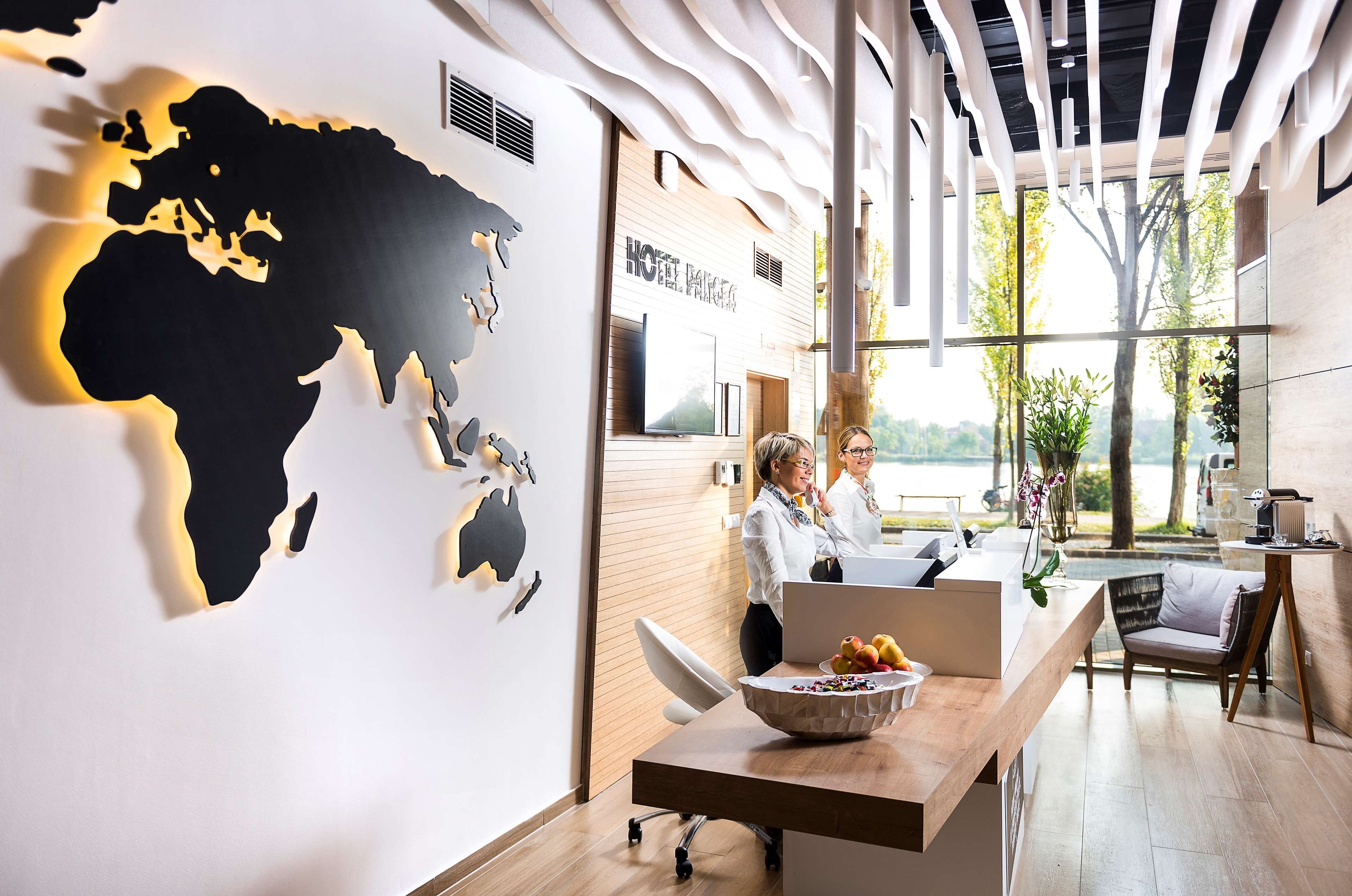 Un área de recepción moderna de hotel con un gran mapa del mundo en la pared, un mostrador de madera y vistas al exterior.
