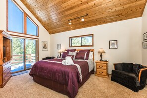 A bedroom with a large bed, wooden ceiling, two windows, a chair, and a nightstand.