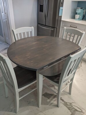 Une table de salle à manger ronde, entourée de quatre chaises blanches, un réfrigérateur en acier inoxydable et une machine à café posée sur un plan de travail blanc.