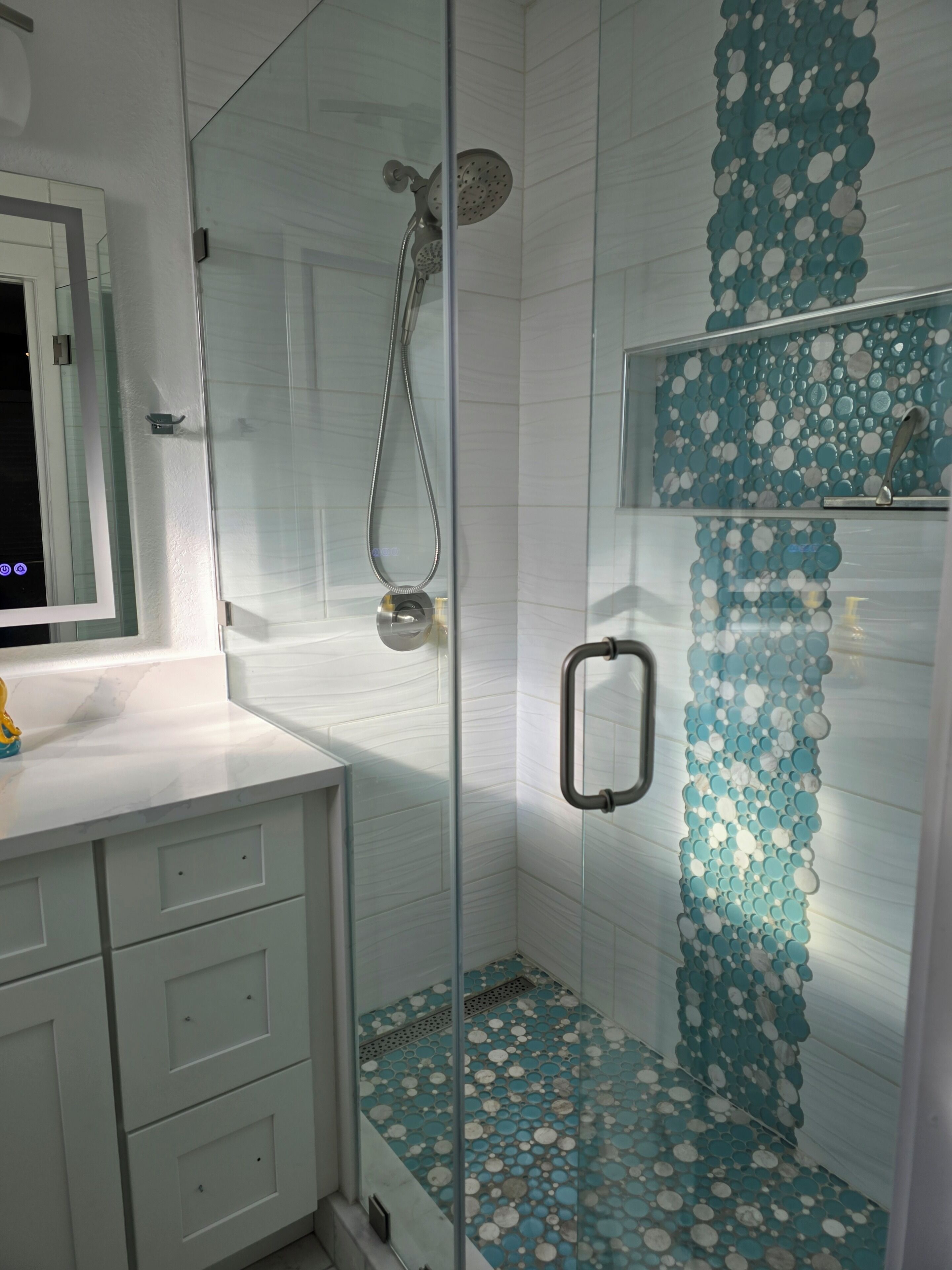 Ducha con mampara de vidrio, cabezal de ducha tipo lluvia, pared de azulejos blancos y diseño decorativo de baldosas azules y blancas en el suelo.