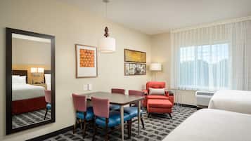 A hotel room with a bed, a dining table with chairs, a red armchair, a large mirror, and artwork on the walls.