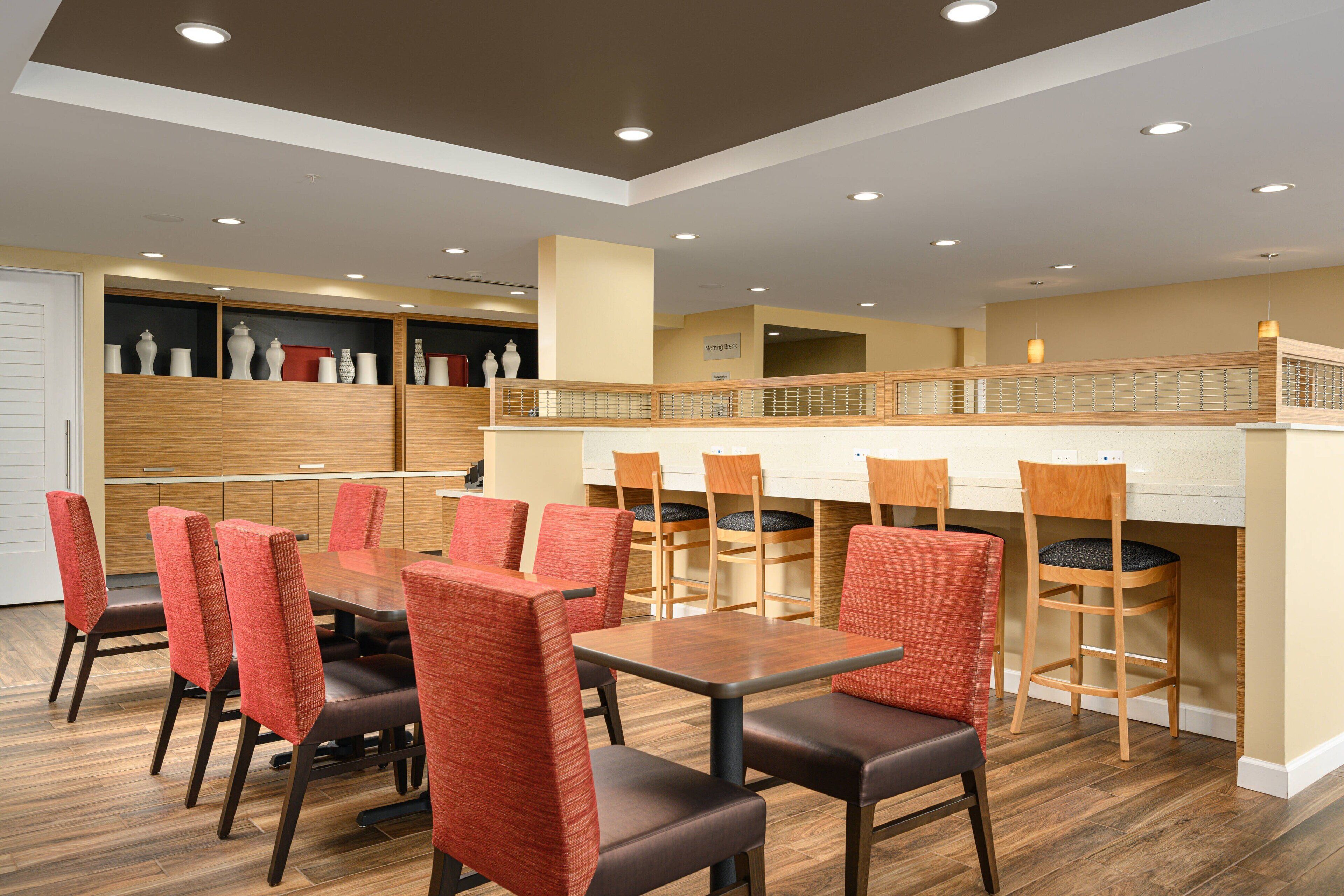 Un espace repas avec des tables en bois et des chaises à revêtement rouge, un coin cuisine avec des meubles en bois, et un balcon doté d’une balustrade.