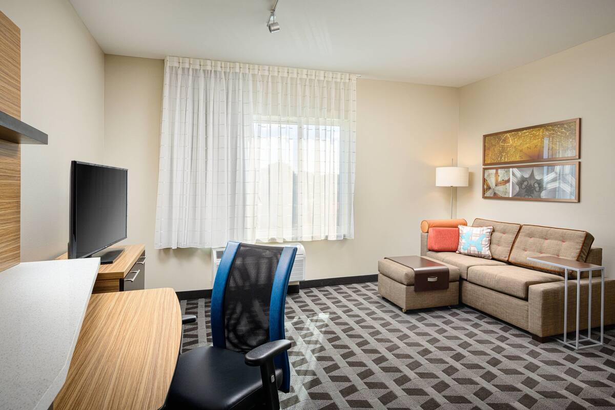 Ein Hotelzimmer mit Flachbildfernseher, Schreibtisch, Stuhl, Sofa, kleinem Tisch, Lampe und einem gemusterten Teppich.