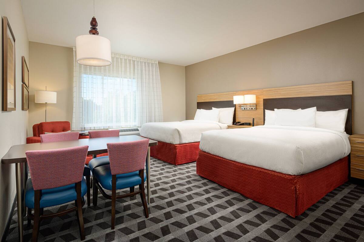 Ein Hotelzimmer mit zwei Betten, einem Esstisch mit Stühlen, einem Schreibtisch und einem großen Fenster mit Vorhängen.