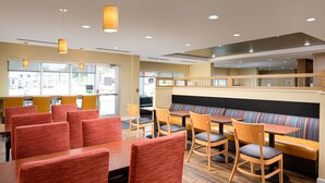 A modern restaurant interior with wooden tables and chairs, a central seating area with a cushioned bench, and large windows providing a view of the outside.