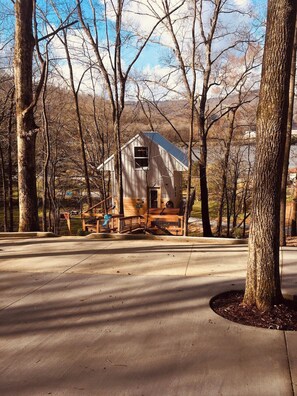 A rustic cabin with a sloped roof, surrounded by trees, and a paved driveway leading to a wooden deck.