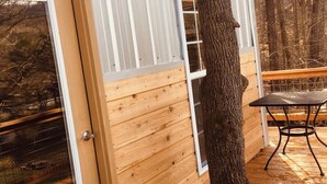 A wooden deck with a metal table and chairs, a door, and a tree trunk. - Blue Heron Treehouse - Private Hot Tub (Guild)