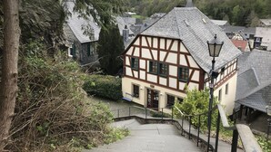 Une maison traditionnelle à colombages, dotée d’un escalier en pierre menant à l’entrée, entourée de verdure et située dans un paysage vallonné.