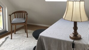 Une pièce avec un plafond en pente, un puits de lumière, une chaise en bois, une table recouverte d’une nappe à rayures et une lampe.