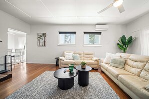 Un salon moderne avec un canapé beige, deux petites tables rondes, une grande plante et un ventilateur de plafond.