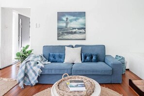 Un salon avec un canapé bleu, une table basse en osier et un tableau encadré représentant un phare, accroché au mur.