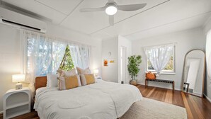 A bedroom with a bed, bedside tables, a ceiling fan, and a window with sheer curtains. - Kings Beach Home Amongst Tranquil Gardens (Kings Beach)