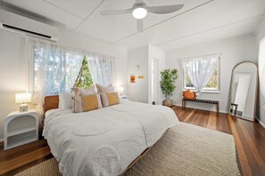 A bedroom with a bed, bedside tables, a ceiling fan, and a window with sheer curtains. - Kings Beach Home Amongst Tranquil Gardens (Kings Beach)