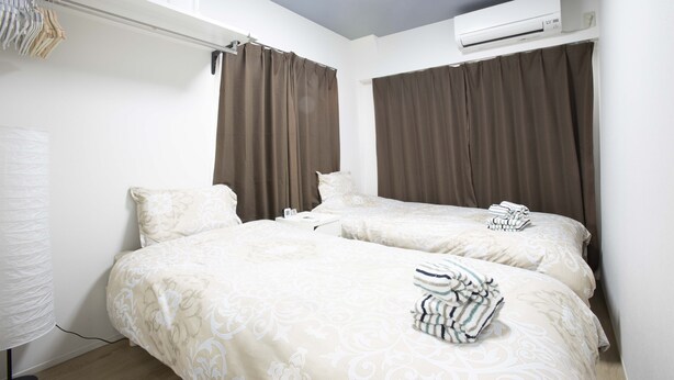 A hotel room with two beds, a ceiling fan, an air conditioning unit, and a window with curtains. - Koenji Street Apartment (Tokyo)